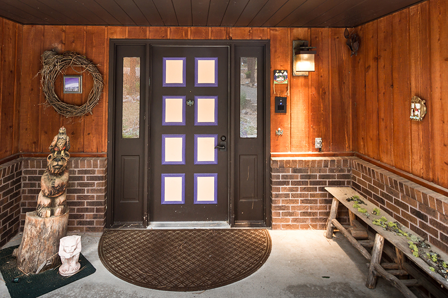 A wide porch with a brown, zero-step entrance door allows a variety of decorations without interfering with the entrance, including a wooden bear statue, a white crouched gargoyle, and a wooden bench covered in plants and sticks.